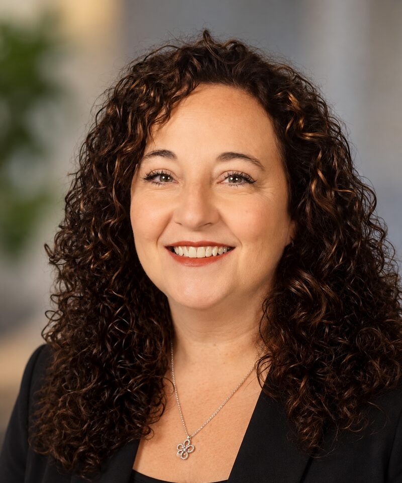 Paula R. Henderson, smiling, with shoulder-length curly dark brown hair and a black blazer.